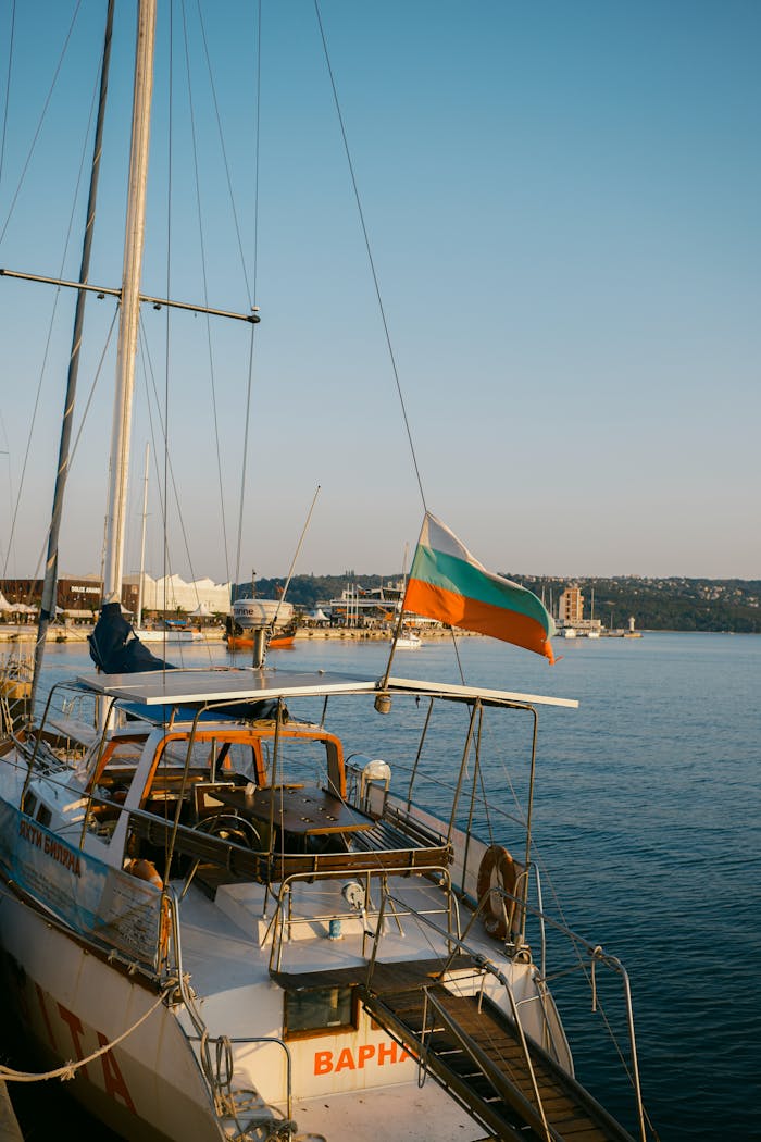 A serene view of a sailboat docked in Varna, Bulgaria, with the national flag flying and the sea reflecting the sunset glow.